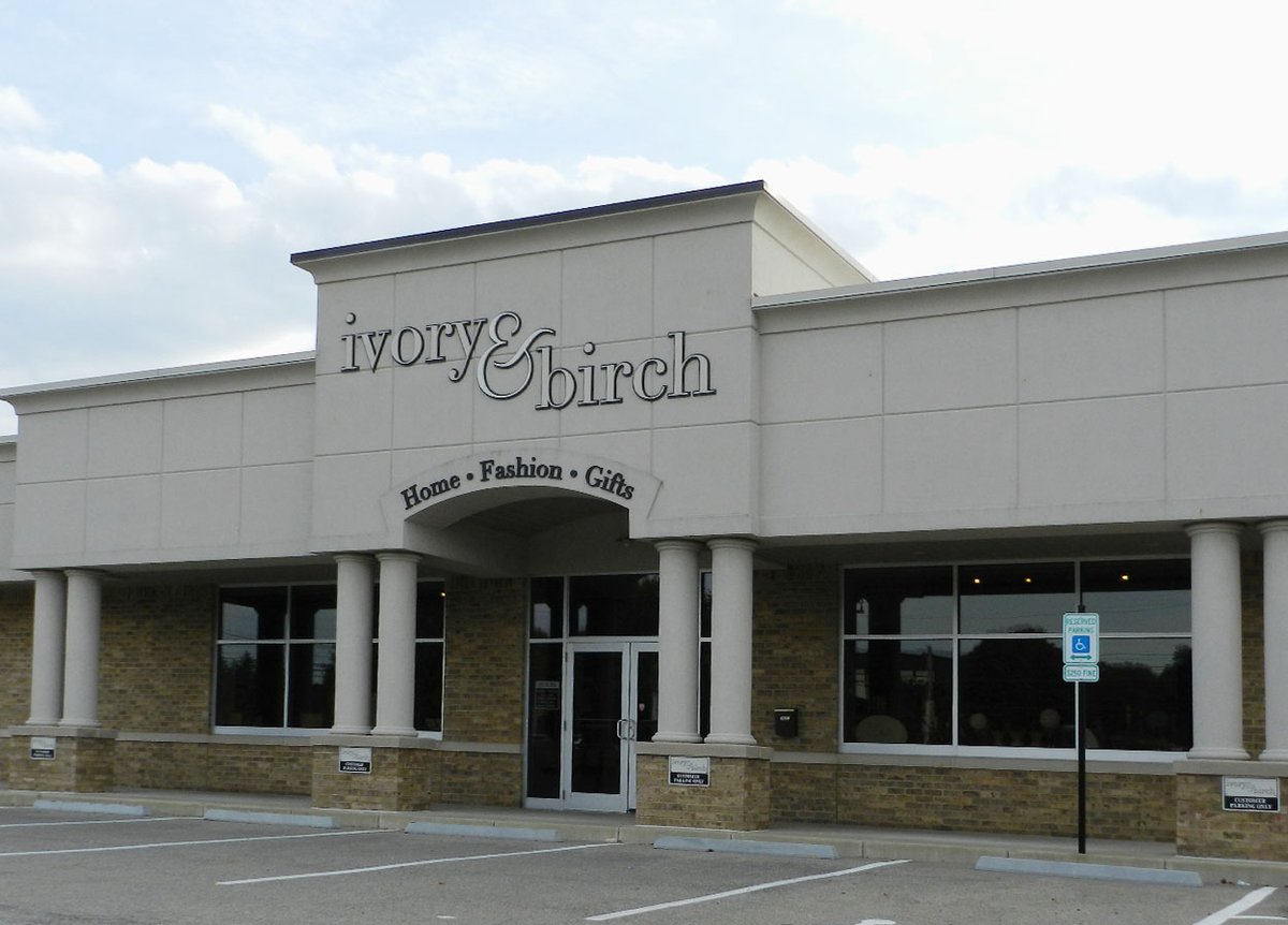 Exterior of the ivory & birch store. The building features a light-colored facade with a sign that reads 'ivory & birch' above the entrance. Below the sign, it says 'Home, Fashion, Gifts.' The entrance is flanked by large windows and two columns. A parking lot with designated accessible parking spaces is in front of the building. The sky is partly cloudy.