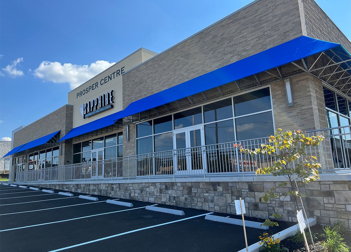 Exterior of the Prosper Centre building with a sign for 'Sapphire Aesthetics and Wellness.' The building is made of light-colored brick and has blue awnings above the windows and doors. There is a metal railing along the front, and the parking lot has freshly painted lines. A small tree is planted in the landscaped area in front of the building. The sky is clear and blue.