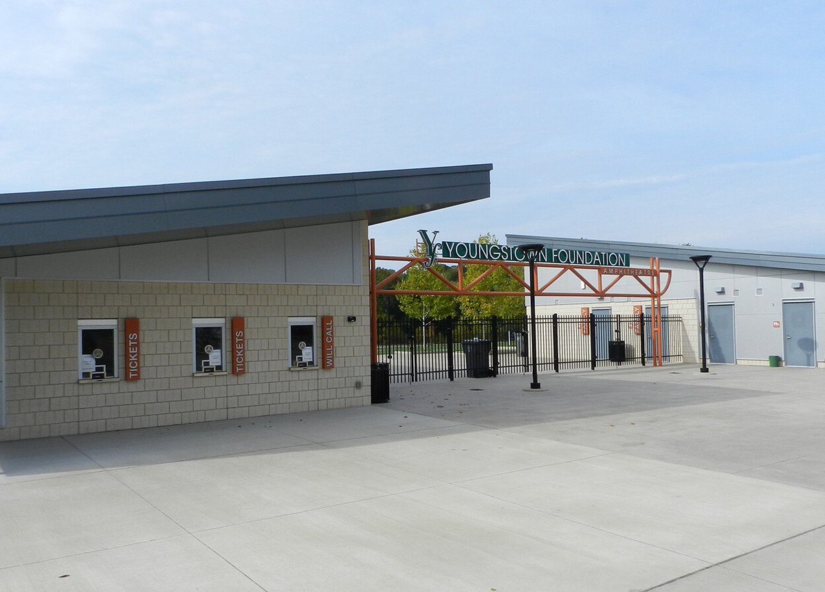 Entrance to the Youngstown Foundation Amphitheatre. The ticket booth, with three windows labeled 'Tickets' and 'Will Call,' is situated under a modern roof. An orange metal framework supports a sign that reads 'Youngstown Foundation Amphitheatre.' The area is paved, and there are some trees visible in the background under a cloudy sky.