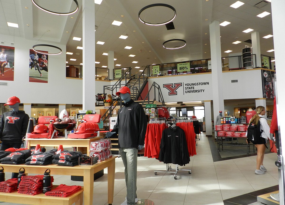 Interior of a Youngstown State University merchandise store. The store features mannequins wearing YSU apparel, tables and racks displaying red and black clothing, and various university-branded items. Large banners and posters decorate the walls, and a staircase leads to an upper level. A sign on the wall reads 'Youngstown State University' with the YSU logo.