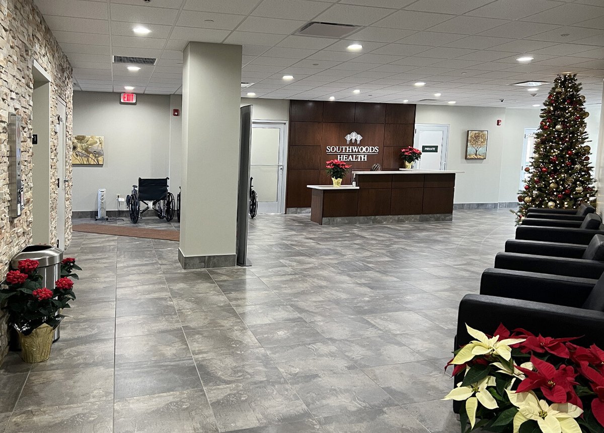 Interior of the Southwoods Health building, featuring a reception desk with a sign that reads 'Southwoods Health.' The lobby has a tiled floor, black chairs, and a large Christmas tree decorated with ornaments. To the left, there are wheelchairs and a stone wall with a piece of artwork. Poinsettia plants are placed throughout the lobby, adding festive decor.