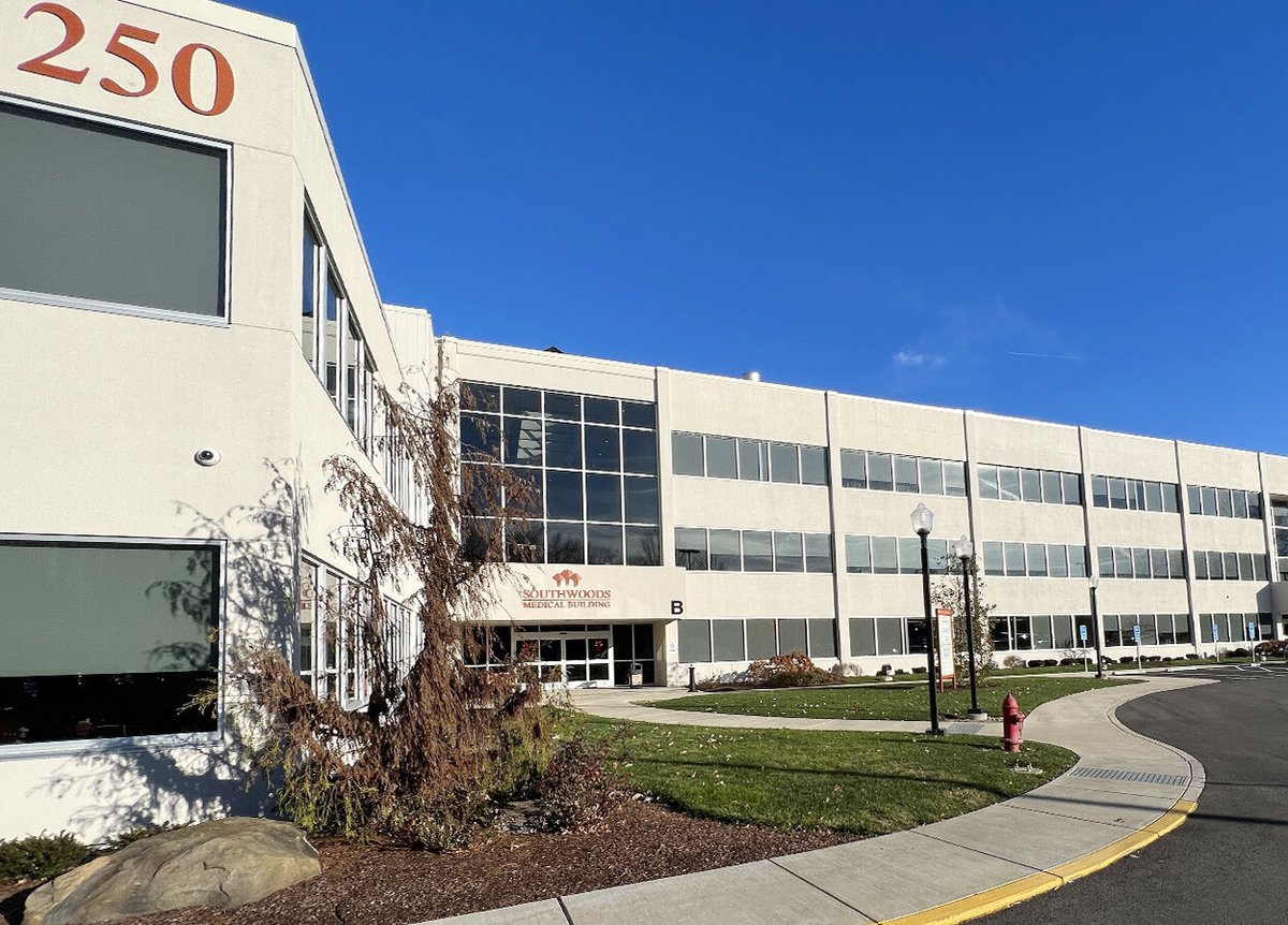 Exterior of the Southwoods Medical Building with a large number '250' on the corner. The building is a modern, multi-story structure with numerous windows. The entrance is marked with a sign reading 'Southwoods Medical Building,' and a letter 'B' is visible near the door. The surrounding area is landscaped with grass, a rock, a tree, and a sidewalk. The sky is clear and blue.