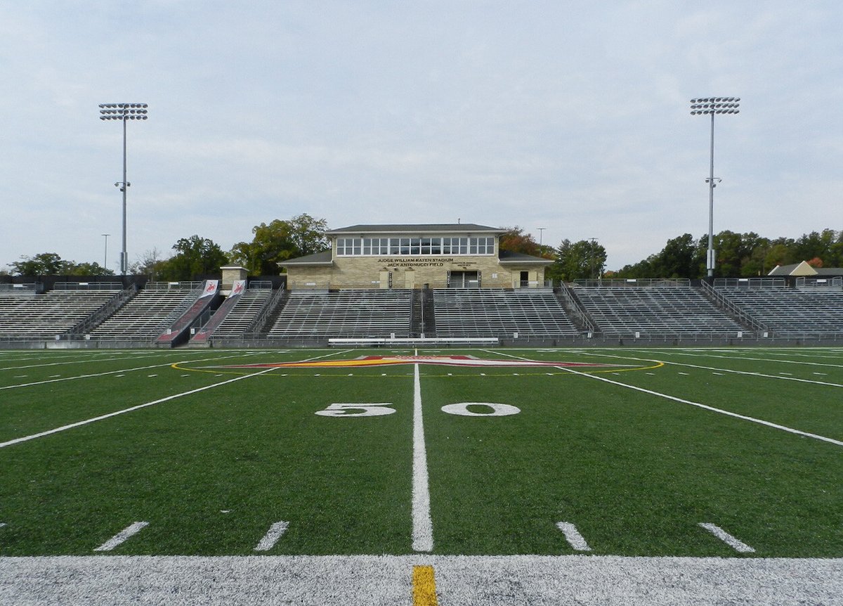 A view of a football stadium from the 50-yard line. The stadium features a central building with a sign that reads 'Judge William Ray Stadium - Nick A. Loparo Sr. Field,' surrounded by bleachers on both sides. The field is green with white yard markers, and the end zones and mid-field feature team logos. Two tall light poles are visible, one on each side of the bleachers, and trees can be seen in the background under a cloudy sky.