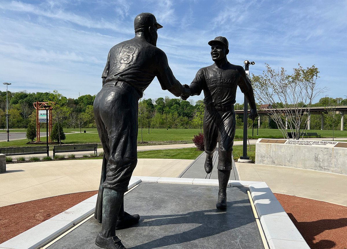 A statue of two baseball players shaking hands on a concrete platform. The player on the left has the number 13 on his back, and both players are dressed in vintage baseball uniforms. The background includes a park with green trees, benches, and a sign that reads 'Wean Park'.