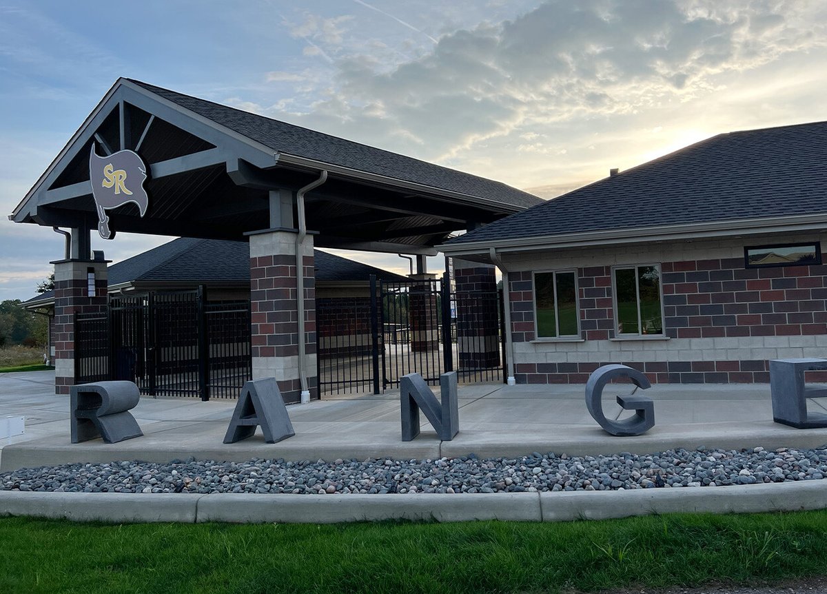 A building with a pitched roof and brick columns. The entrance is gated, and above it is a sign with a flag featuring the initials 'SR.' In the foreground, large concrete letters spelling 'RANGE' are positioned on a bed of rocks. The sky is cloudy, and the grass is green.