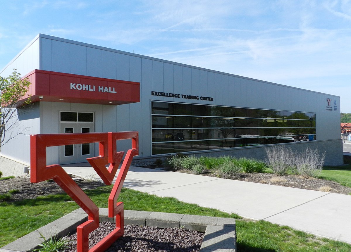 A modern building with a red sign that reads 'Kohli Hall' and a large window with the words 'Excellence Training Center' above it. In the foreground, there is a large red sculpture of the letter 'Y' on a bed of rocks. The building is surrounded by a well-maintained lawn and landscaping, and the sky is clear and blue.