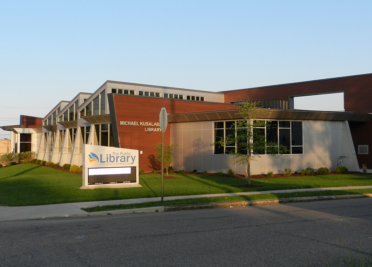 Exterior of the Michael Kusalaba Library, a modern building with large windows and a mix of wood and metal paneling. A sign in front of the building reads 'The Public Library of Youngstown & Mahoning County.' The surrounding area is landscaped with grass, small trees, and shrubs. The sky is clear and blue.