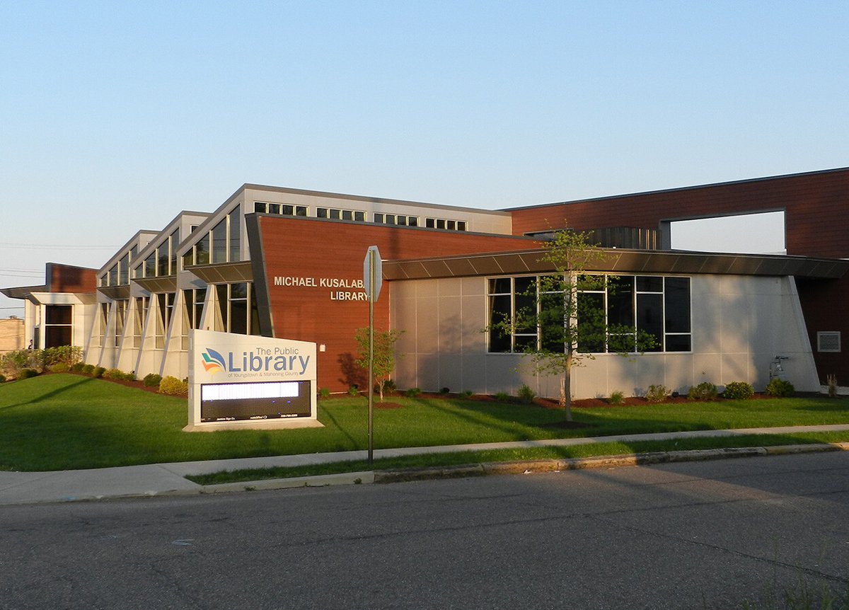 Exterior of the Michael Kusalaba Library, a modern building with large windows and a mix of wood and metal paneling. A sign in front of the building reads 'The Public Library of Youngstown & Mahoning County.' The surrounding area is landscaped with grass, small trees, and shrubs. The sky is clear and blue.