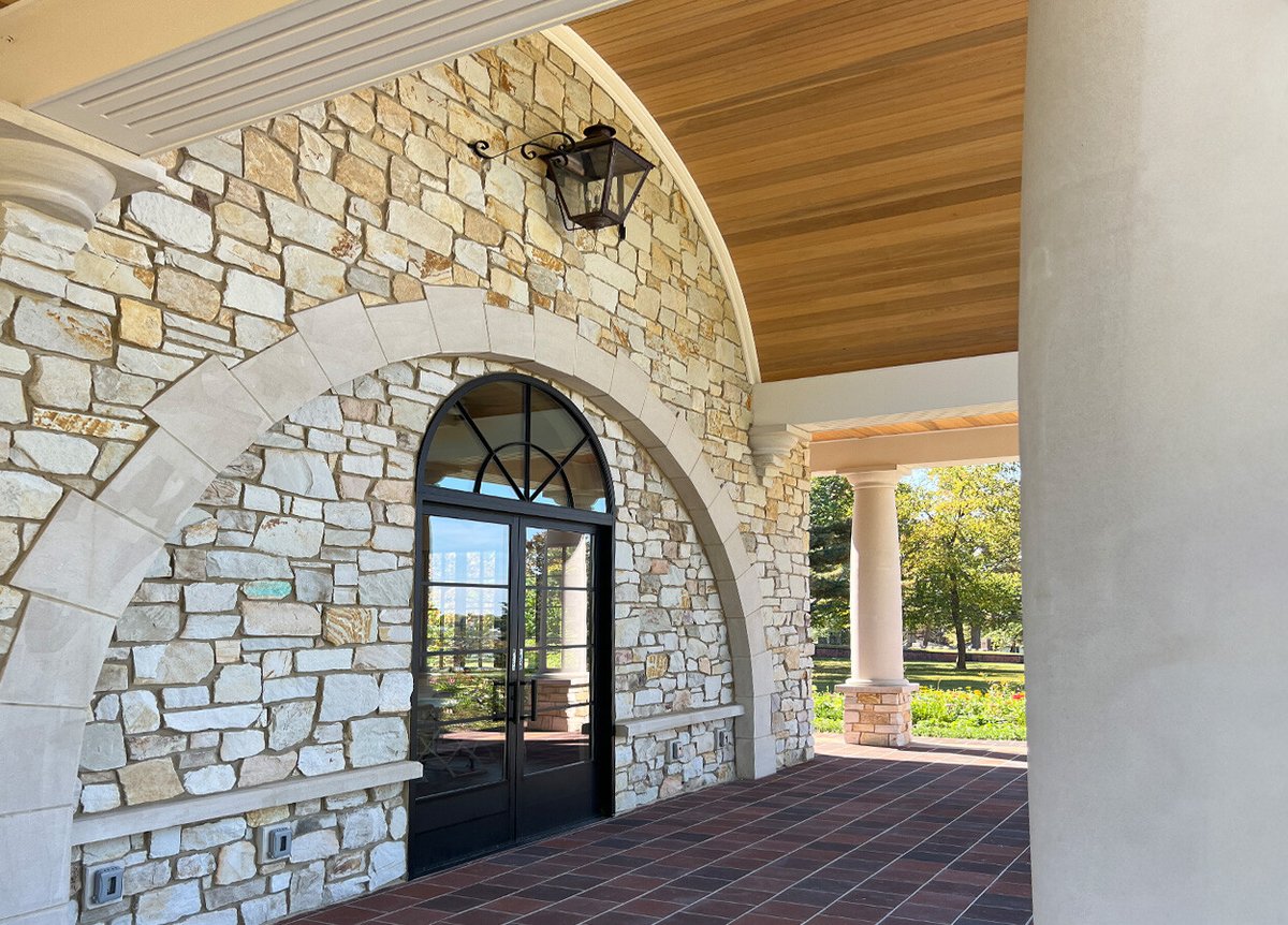 An outdoor area with a stone archway and columns, featuring a glass door with a semi-circular window above. The ceiling is wooden, and the ground is covered with reddish-brown tiles. The background shows a garden with green trees and bushes.