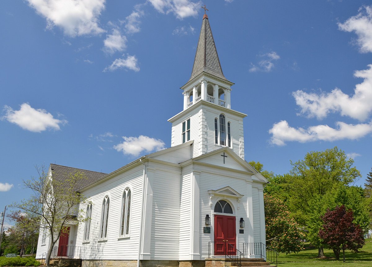 An exterior view of a white church with a tall steeple and red double doors. The church features arched windows and is set against a clear blue sky with scattered white clouds. The surrounding area includes green trees and a well-maintained lawn.