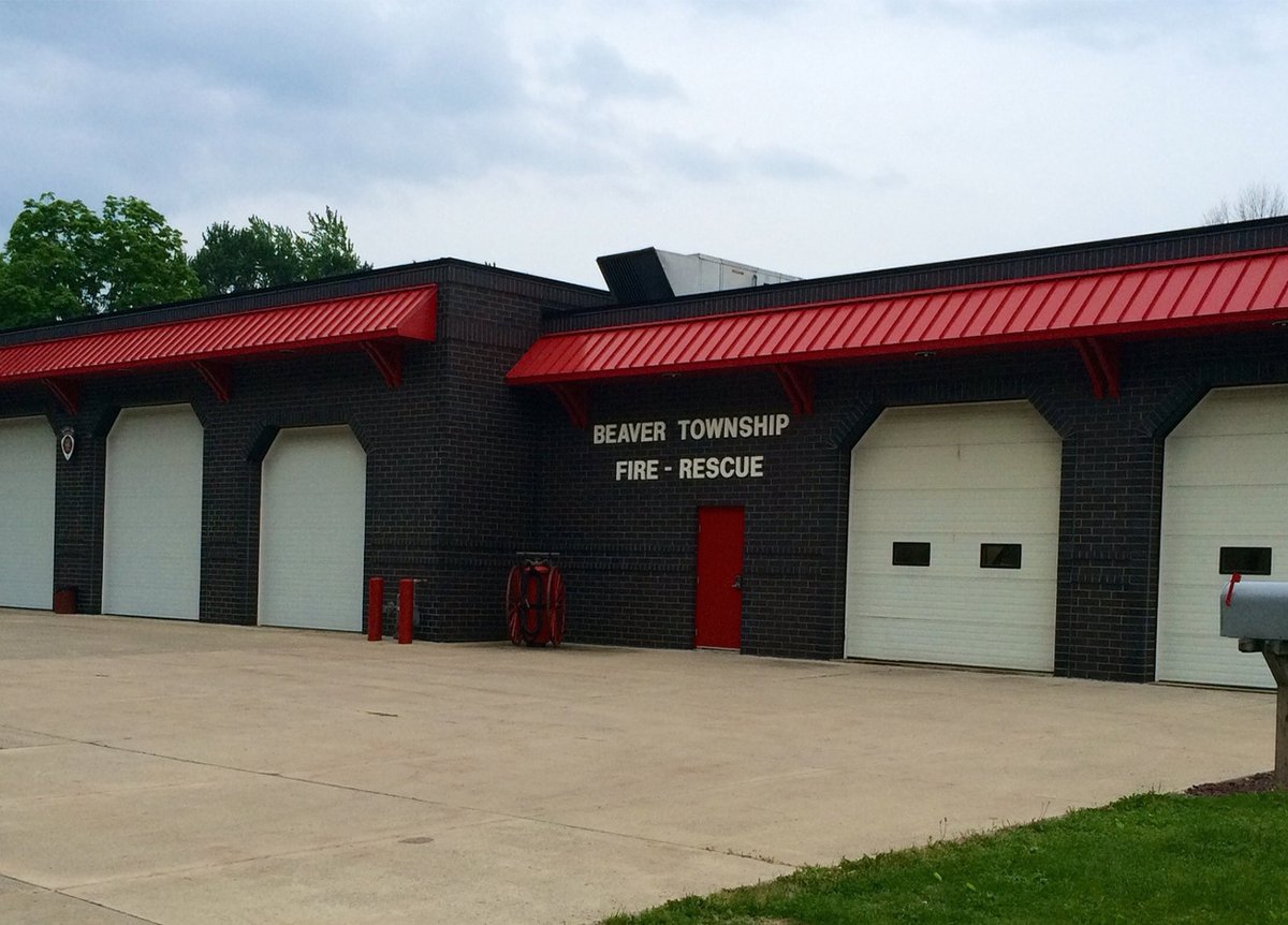 Exterior of the Beaver Township Fire-Rescue building. The structure is made of dark bricks with red metal awnings above the garage doors and the entrance. There are three white garage doors and a red entry door. The sign on the building reads 'Beaver Township Fire-Rescue.' The area in front is paved, and the sky is overcast.